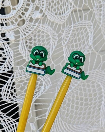 Pair of yellow knitting needles with green silicone bookworm-shaped stoppers perched on books, set against a delicate white lace background