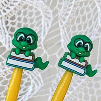 Close-up of two yellow knitting needles with green bookworm-shaped silicone stoppers resting on a white lace backdrop.