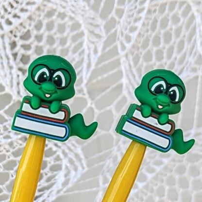 Close-up of two yellow knitting needles with green bookworm-shaped silicone stoppers resting on a white lace backdrop.