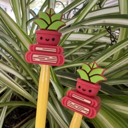 Two silicone needle stoppers shaped like red pots with smiling red and green succulent plants sitting on top of two red books. The stoppers are placed on yellow knitting needles. Green and white striped plant leaves fill the background.