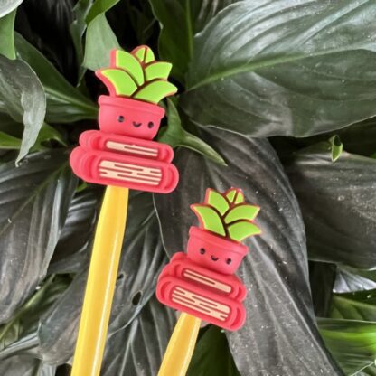 Two silicone needle stoppers shaped like red pots with smiling red and green succulent plants sitting on top of two red books. The stoppers are placed on yellow knitting needles. Dark green plant leaves fill the background.